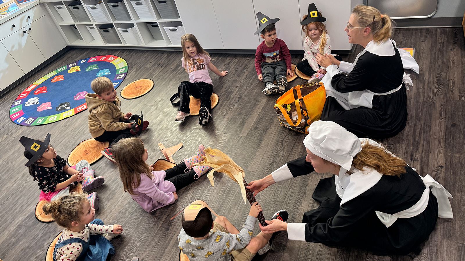 Preschool children sit in a circle listening to teachers during a themed lesson.