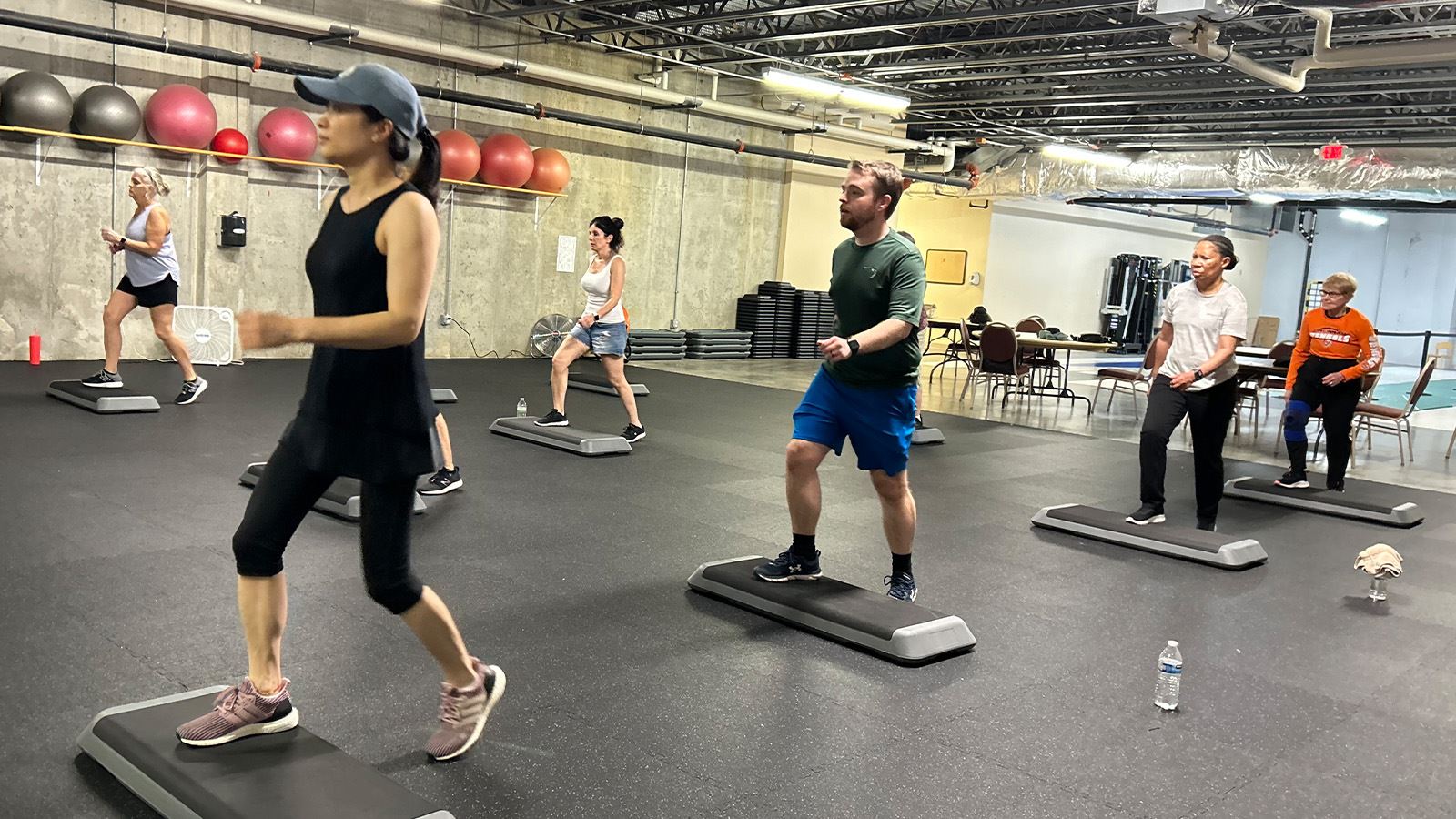 Adults take part in a step aerobics class in a group fitness room.