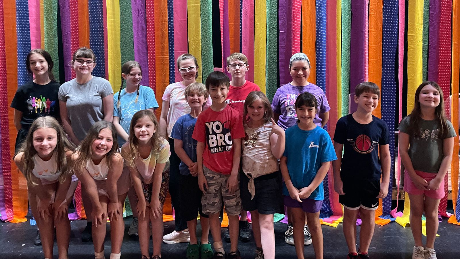 Group of smiling children standing on stage in front of a colorful fabric backdrop.
